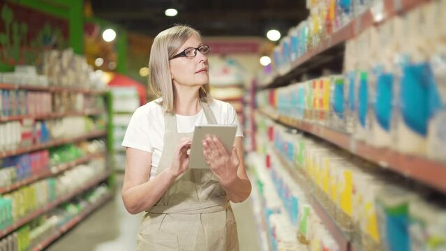 Senior female manager worker in supermarket, grocery store or deli in apron with digital tablet. Mature female small business owner employee inspects shelves inventory products doing checking stock