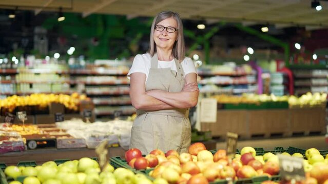 Portrait senior mature woman worker in a Vegetable section supermarket standing smiling with arms crossed. Friendly pleasant female looking at camera in a fruit shop market. Employee greengrocer
