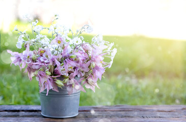 A bouquet of beautiful flowers in an iron bucket on a wooden board and a green background with sunbeams and highlights. Floral background with blurry lights and copy space.