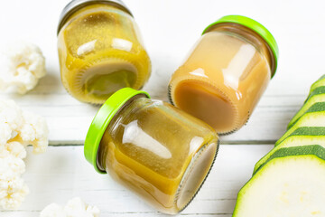 Glass jars with children's mashed broccoli, cauliflower and zucchini puree on a white wooden background, side view. Baby food. The first lure of the baby.