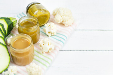 Obraz na płótnie Canvas Glass jars with children's mashed cauliflower and zucchini puree on a white wooden background. Space for the text. Baby food. The first lure of the baby.