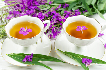 Obraz na płótnie Canvas A drink (infusion, decoction) of ivan-tea in a white cup on a saucer on a wooden background. Grass and flowers of the plant kipreya, epilobium, ivan-grass.