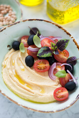 Bowl of hummus with baby tomatoes, black olives, red onion and basil leaves, vertical shot, close-up