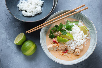 Bowl of thai red chicken curry with coconut milk and white rice, high angle view on a grey concrete background