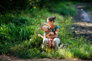 Mom and son in park among grass are playing fishing with stick and fishing rod. Mom hugs boy, helps him. concept of happy childhood in nature and happy single parent