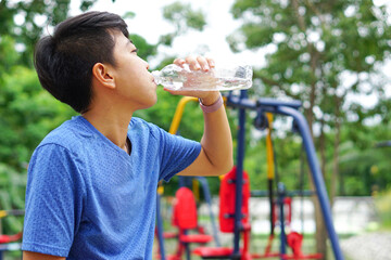 Asian young man drinking water after jogging, an outdoor exercise machine on the back. For good health, loss weight, good shape.
