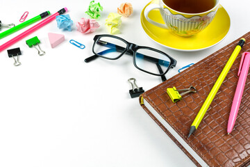 Business concept. A brown notebook, a yellow cup of tea on a saucer, black glasses, a pencil, a pink pen, colored paper clips on a white background. Space for text. Freelance. Office worker's desktop.