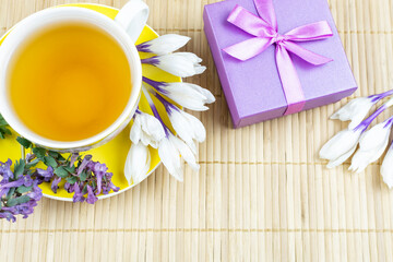 A yellow cup of black tea on a saucer, spring flowers crocuses and tufts and a box with a gift and a bow on a wooden bamboo background. Space for the text. The concept of the holiday. Good morning.