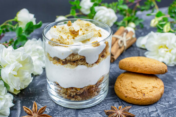Multi-layer cottage cheese banana dessert with oatmeal cookies and yogurt, cinnamon sticks, anise and bushy white roses on a black (dark) background. Close-up. Romantic breakfast.