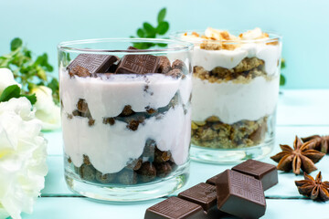 Multi-layer cottage cheese dessert with chocolate balls, yogurt and chocolate slices and banana dessert in a glass glass, white bush roses and anise on a blue (beryuzov) background. 