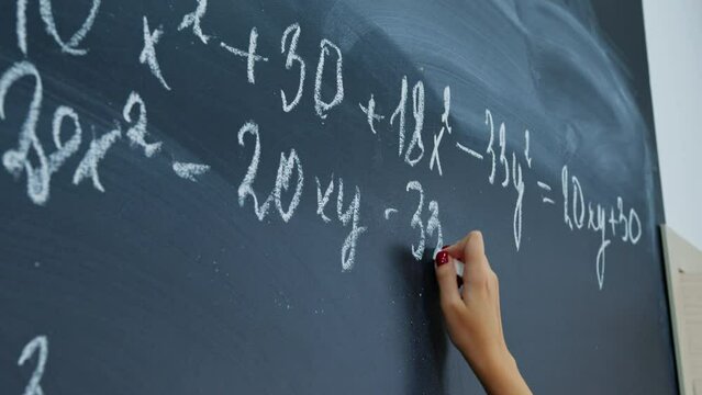 Female hand with chalk. Math faceless teacher writes examples for schoolchildren on blackboard. Student, pupil girl solves system of equations on chalkboard. Algebra, mathematics in classroom