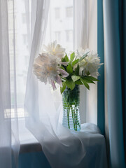 A bouquet of peonies in a transparent striped vase on the windowsill by the window. Blue curtains...