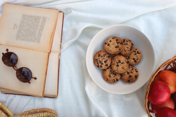 Picninc blanket with straw bag, bowl of strawberries and blueberries, bowl of chocolate chip cookies, books, sunglasses and basket of apples in the garden. Top view.
