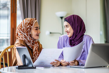 Two Asian muslim women wearing hijab working together with using laptop pc tablet and paper showing...