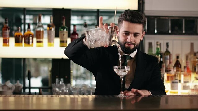 Caucasian handsome bartender pouring cold refreshing drink into glass through strainer. Bearded young man wearing black suit and tie at work of luxury restaurant.