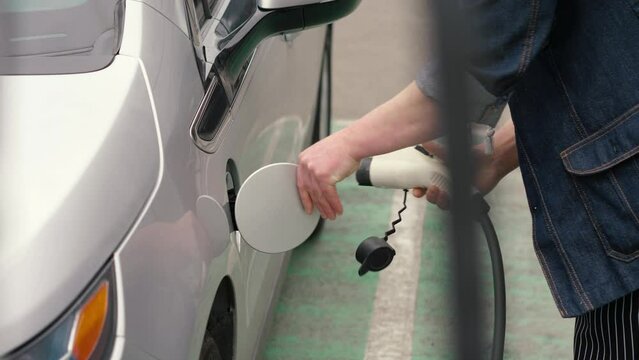 Male hand plugging in charging cable to electric vehicle and charges batteries. EV station in parking lot. Unrecognizable man inserts power connector into EV car.