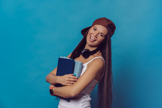 Young brunette college girl with long hair dressed in white t-shirt and baseball cap, broad smiling, holds her school books and notebooks, with headphones around her neck, over blue backdrop.