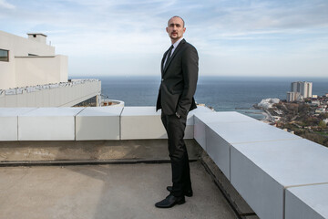 a man in a black suit and tie stands on a roof with the sea skyline in the background