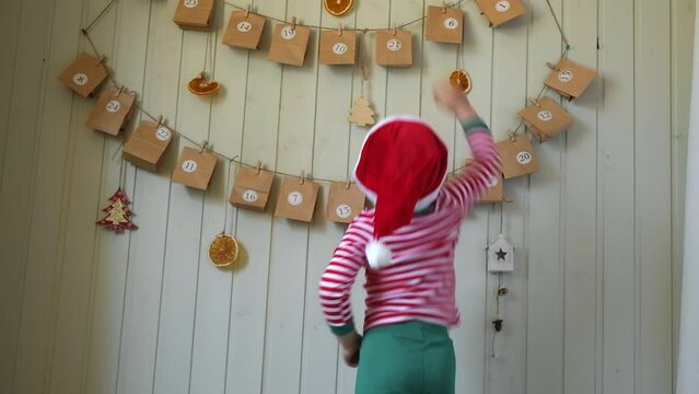 Boy in pajamas, Santa cap stretches for paper advent calendar with presents. Little happy curious child opening gift of handmade Christmas calendar hanging on wall. Celebrating at home, Xmas tradition