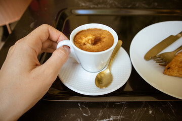 Small ceramic or porcelain white cup with hot coffee drink - espresso, cappuccino, mochaccino - in a woman's hand. Breakfast, brunch in a cafe, restaurant. Coffee cup on a tray on a black marble table