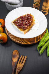 Tostada de jamon con tomate, aceite y pimienta sobre un plato blanco colocado encima de una superficie de madera rustica junto a utensilios de cocina, salero e ingredientes de la receta como tomate