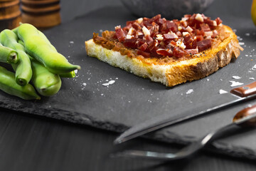 Ham toast with tomato, oil and pepper on a slate plate placed next to rustic wooden kitchen utensils, salt shaker and recipe ingredients such as tomato and broad beans