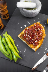 Ham toast with tomato, oil and pepper on a slate plate placed next to rustic wooden kitchen utensils, salt shaker and recipe ingredients such as tomato and broad beans