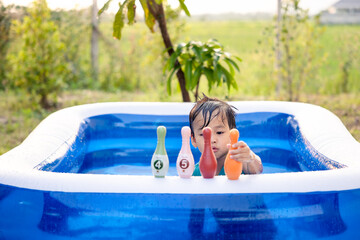 Portrait of asian child, kid or little boy to play plastic bowling toy in blue portable inflatable swimming pool at outdoor, backyard or garden in summer evening. Include green field, sky background. - Powered by Adobe