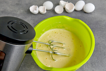Beaten eggs with sugar in a green bowl with a beater immersed in a mixer and eggshells from chicken eggs