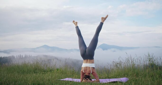 Sporty female athlete in activewear doing headstand exercise on yoga mat outdoors. Young woman spending leisure time on nature for active workout.