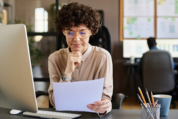 Young manager in eyeglasses sitting at her workplace in front of computer and reading contract with...