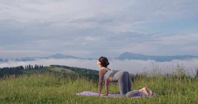 Active caucasian woman in sport clothes standing on yoga mat in downward-facing dog pose. Young lady having workout among summer nature of mountains.