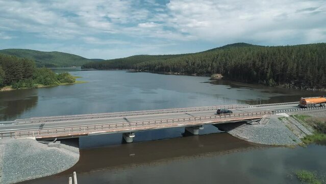 Aerial view of small road bridge across the river in rural area. Cars driving on the bridge. Sky reflections on the river water surface