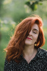 Portrait of gorgeous middle aged woman in casual dress relaxing in a blooming spring garden. Happy senior woman smiling and looking at camera. Red-haired mature lady posing outdoors in a spring day.