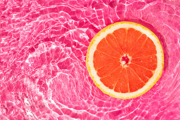 Fresh round grapefruit slice floating in a pool with a vivid pink water. Minimal summer fruit creative art. Aesthetic food concept.