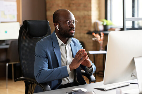 African confident businessman in suit sitting on chair at his workplace in front of computer monitor and talking to colleague online