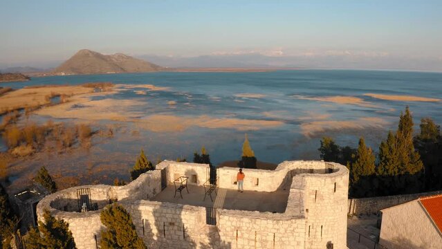 Young woman in modern clothes walks to the edge of the Besac Fortress in Montenegro with fantastic view over Skadarsko Jezero, which is partly lit by the setting sun. Backwards drone dolley tilt shot