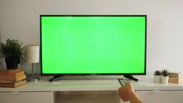 Woman watching TV with green screen switching channels, using remote control in living room.