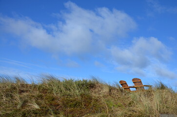 Two empty deckchairs on sand dunes overgrown with grass, blue sky with clouds background
