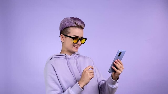 Joyful young adult woman with short dyed purple hair wearing violet sweatshirt, yellow sunglasses, smiling, surprising to find funny memo, laughing, using smartphone isolated on monocolor background