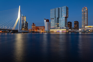 Rotterdam nighttime panorama with “Erasmus-Bridge“ over river Nieuwe Maas at evening blue hour in South Holland Netherlands. Waterfront with illuminated bridge and tall buildings on the waterfront.