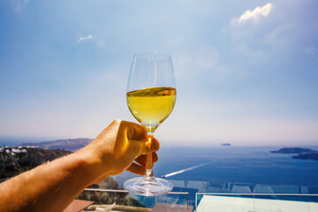 A white wine glass in hand against the backdrop of the Aegean sea and caldera on Santorini island.