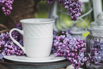 A beautiful postcard. A white coffee cup with a saucer, candles and a vase with a bouquet of purple lilac. The color 2022 is very peri. Beautiful still life. Spring time. The concept of "Good morning"