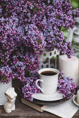 A beautiful postcard. A white coffee cup with a saucer, candles and a vase with a bouquet of purple lilac. The color 2022 is very peri. Beautiful still life. Spring time. The concept of 