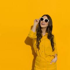 Young woman in linen shirt and shorts posing in sunglasses against yellow wall.
