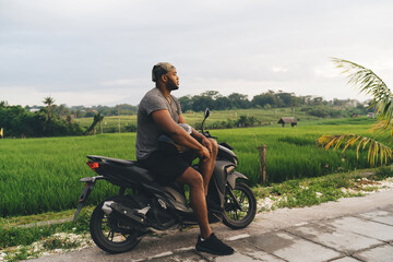 Fototapeta na wymiar Contemplative male tourist with helmet resting at motorbike thinking about driving trip during summer vacations in Indonesia, African American hipster guy in bandanna feeling pondering on rent details