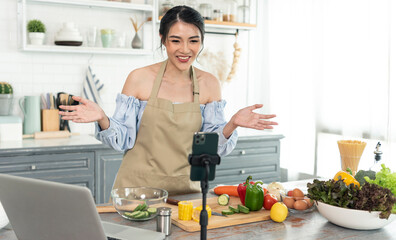 Asian woman food blogger cooking salad in front of smartphone camera while recording vlog video and live streaming at home in kitchen.