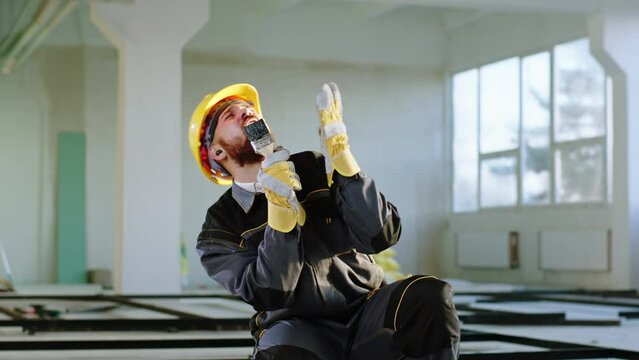 Excited man construction worker at construction site listen music from the wireless earphones and take the painting brush to sing very funny and happy he wearing safety helmet and uniform