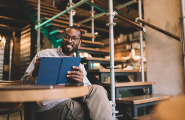 Portrait of happy student in eyewear holding copybook for creating education essay story, cheerful African American male with notebook for organization - learning and studying in coworking space