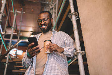 Fototapeta na wymiar Happy hipster guy in optical spectacles using cellular gadget for network socializing during free time, smiling African American man in messaging via mobile application on smartphone device